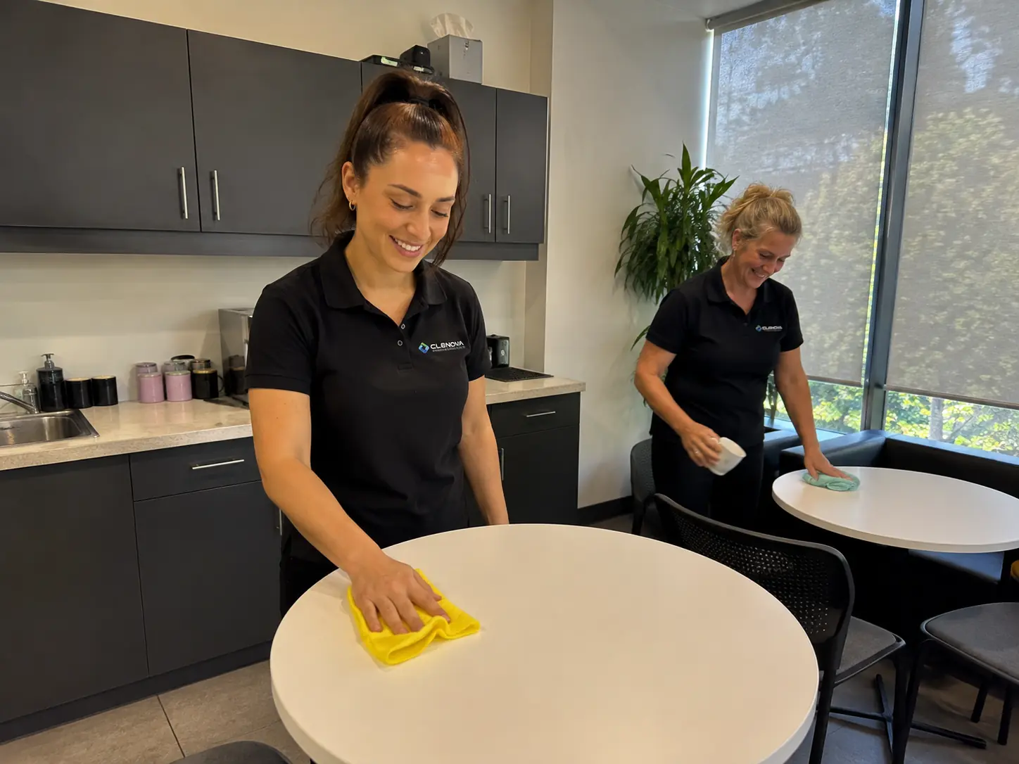Two female office cleaners wiping staff kitchen tables - Illustrative Image